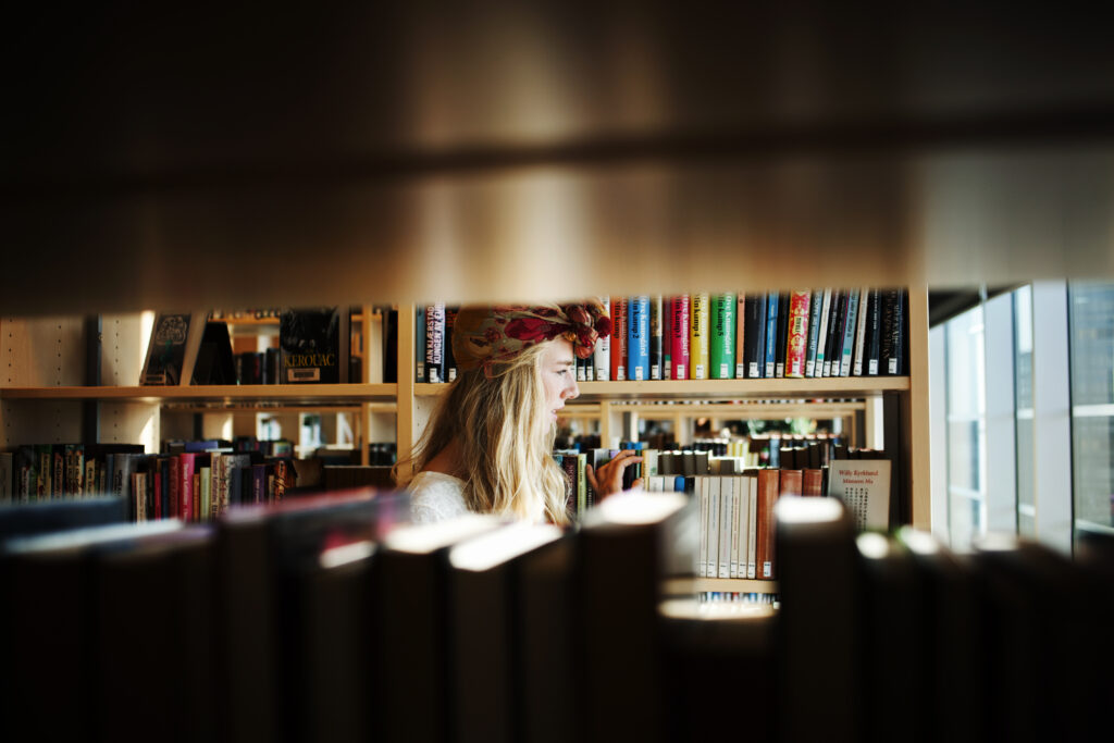 A young woman with wavy blonde hair and a floral headband browses the shelves in a brightly lit library. The shelves are packed with colorful books. The composition is a medium shot, taken at eye level, showcasing the subject's profile.