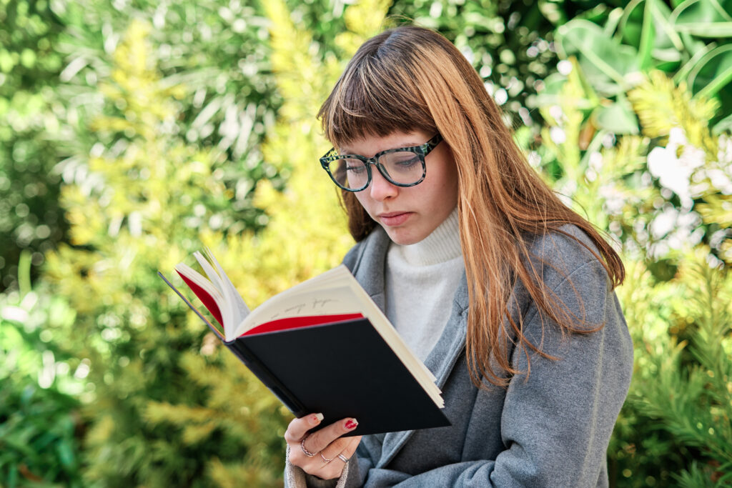 A young woman with long straight brown hair and glasses sits outdoors surrounded by green foliage, absorbed in a book. She wears a grey coat over a white turtleneck. The lighting is bright and soft, enhancing the peaceful, reflective mood.